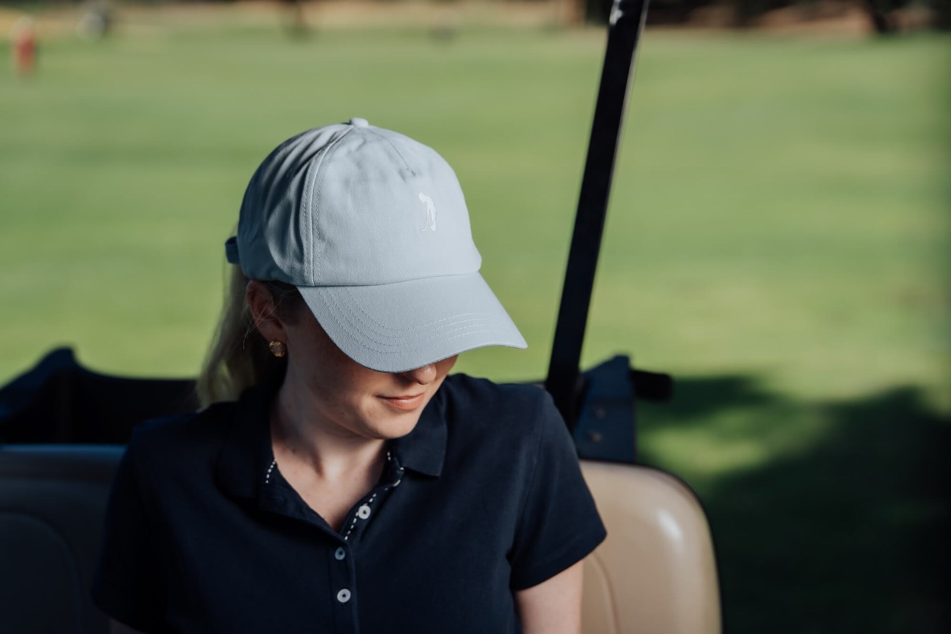 Woman wearing a light blue cap and dark polo shirt sitting in a golf cart on a golf course.