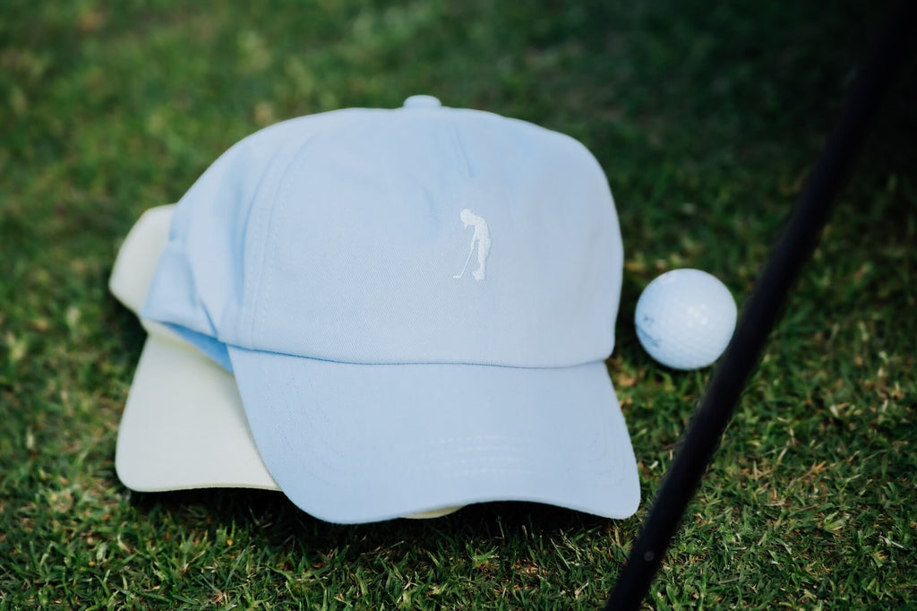 Light blue cap with a logo on a light yellow cap next to a golf ball and club on grass.